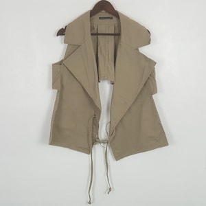May include: A beige sleeveless vest featuring a wide notched collar, open sides, and a drawstring tie closure at the front waist. The garment is constructed with structured lapels and hangs from a dark brown wooden hanger against a plain background.