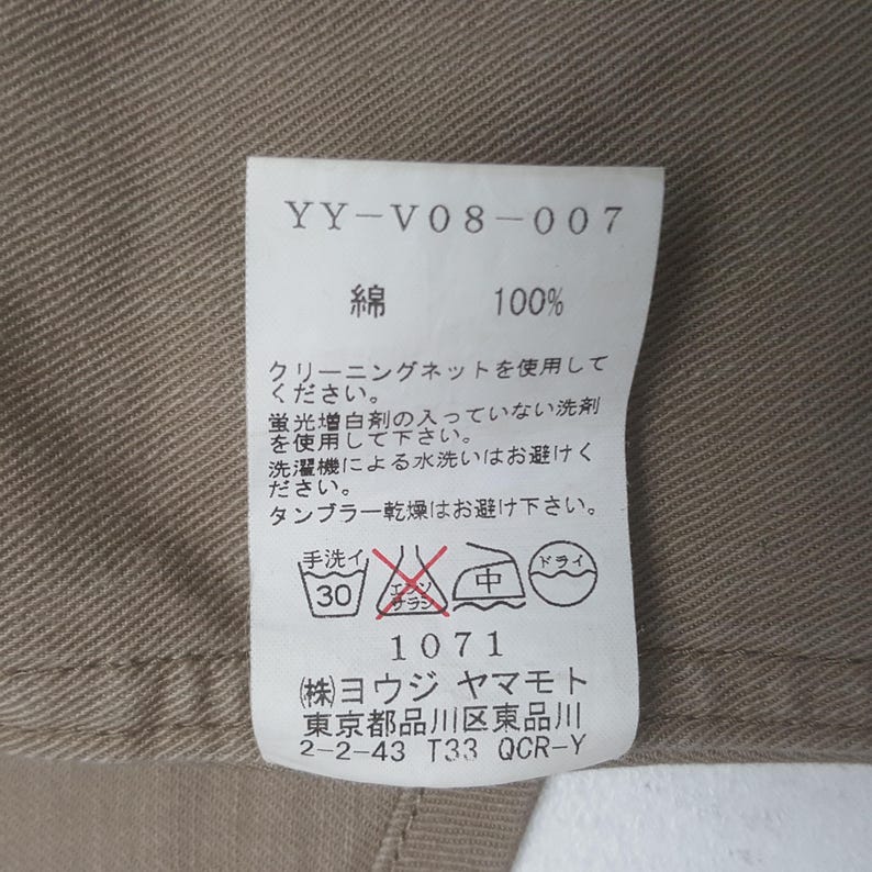 May include: A white rectangular garment care label sewn onto a tan fabric background. The label features the text YY-V08-007, 100% cotton in Japanese, various Japanese care instructions, laundry symbols, and the manufacturer name Yohji Yamamoto.