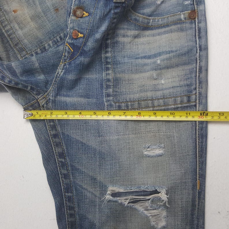 May include: Close-up of distressed blue denim jeans with a measuring tape showing a length of approximately 12 inches. The jeans have a button fly, multiple pockets, and a ripped knee. The denim has a faded, worn appearance.