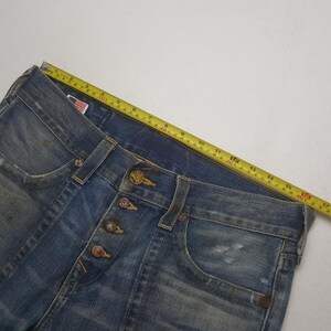 May include: Close-up of blue denim jeans with a button fly. The jeans have a classic five-pocket design and are distressed with faded areas. A yellow measuring tape is laid across the jeans, showing measurements in inches.