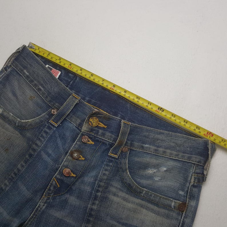 May include: Close-up of blue denim jeans with a button fly. The jeans have a classic five-pocket design and are distressed with faded areas. A yellow measuring tape is laid across the jeans, showing measurements in inches.