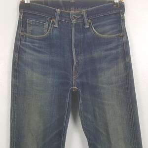 May include: A pair of dark blue denim jeans featuring a high-waisted design, a button fly closure, and copper-toned rivets at the pocket corners. The fabric displays a faded wash pattern with whiskering details around the hips and thighs.