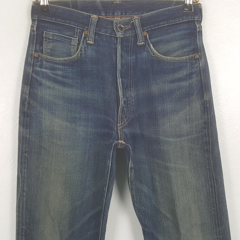 May include: A pair of dark blue denim jeans featuring a high-waisted design, a button fly closure, and copper-toned rivets at the pocket corners. The fabric displays a faded wash pattern with whiskering details around the hips and thighs.