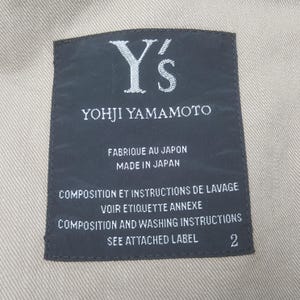 May include: A rectangular black clothing label with white text stitched onto a beige fabric. The label reads Y's YOHJI YAMAMOTO, FABRIQUE AU JAPON MADE IN JAPAN, COMPOSITION ET INSTRUCTIONS DE LAVAGE VOIR ETIQUETTE ANNEXE, and COMPOSITION AND WASHING INSTRUCTIONS SEE ATTACHED LABEL 2.