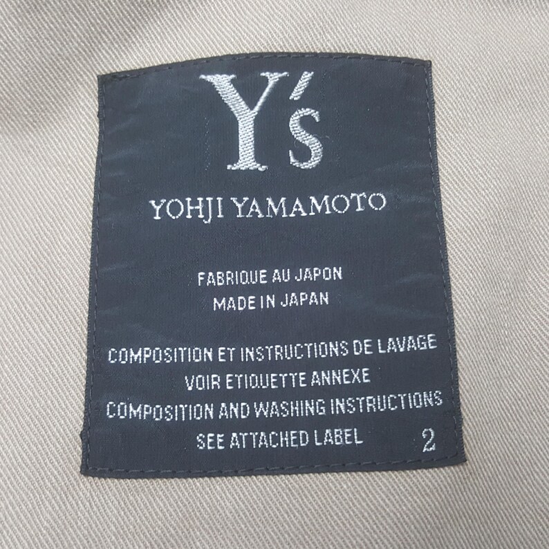May include: A rectangular black clothing label with white text stitched onto a beige fabric. The label reads Y's YOHJI YAMAMOTO, FABRIQUE AU JAPON MADE IN JAPAN, COMPOSITION ET INSTRUCTIONS DE LAVAGE VOIR ETIQUETTE ANNEXE, and COMPOSITION AND WASHING INSTRUCTIONS SEE ATTACHED LABEL 2.