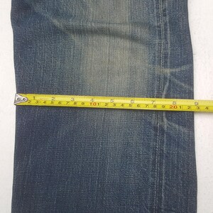 May include: A pair of blue denim jeans featuring a faded wash and visible stitching along the side seam. A yellow measuring tape is placed horizontally across the fabric, indicating a width measurement of approximately 9.25 inches.