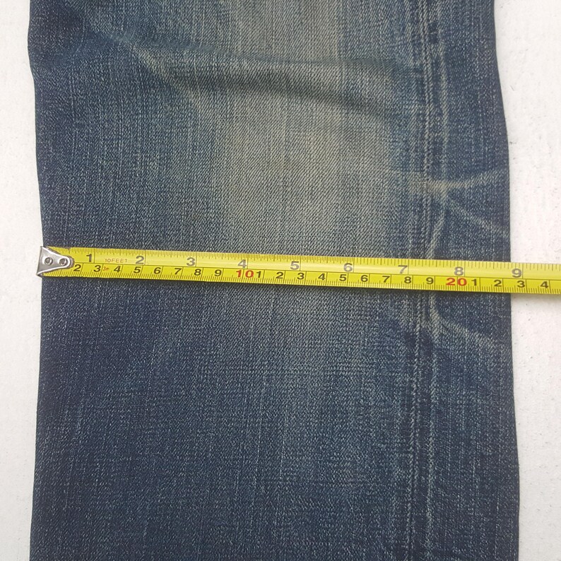 May include: A pair of blue denim jeans featuring a faded wash and visible stitching along the side seam. A yellow measuring tape is placed horizontally across the fabric, indicating a width measurement of approximately 9.25 inches.