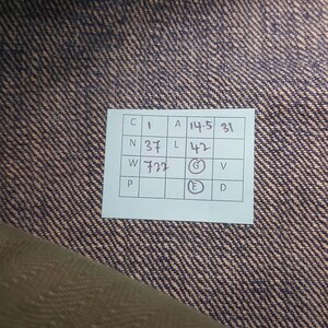 May include: A rectangular white paper tag with a grid containing handwritten text and numbers, including C 1, A 14.5, 31, N 37, L 42, W 722, G, V, P, E, and D, placed on a dark blue and tan diagonal twill fabric with a visible woven texture pattern.