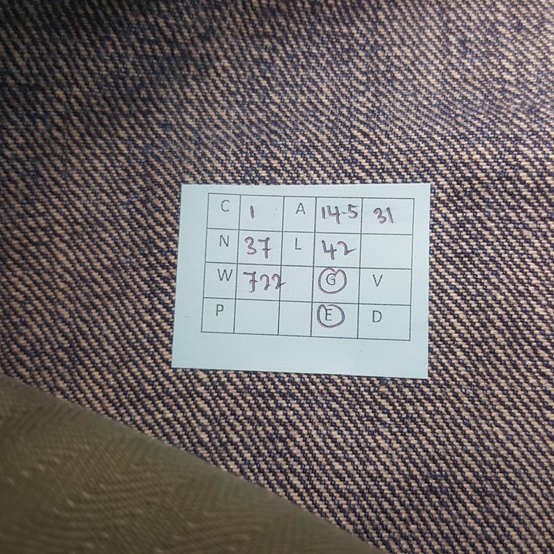 May include: A rectangular white paper tag with a grid containing handwritten text and numbers, including C 1, A 14.5, 31, N 37, L 42, W 722, G, V, P, E, and D, placed on a dark blue and tan diagonal twill fabric with a visible woven texture pattern.
