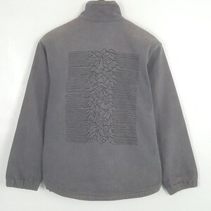May include: A grey long-sleeve jacket featuring a high collar and a black graphic print on the back. The print displays a series of horizontal lines forming a mountain-like wave pattern across the center of the back panel of the garment.