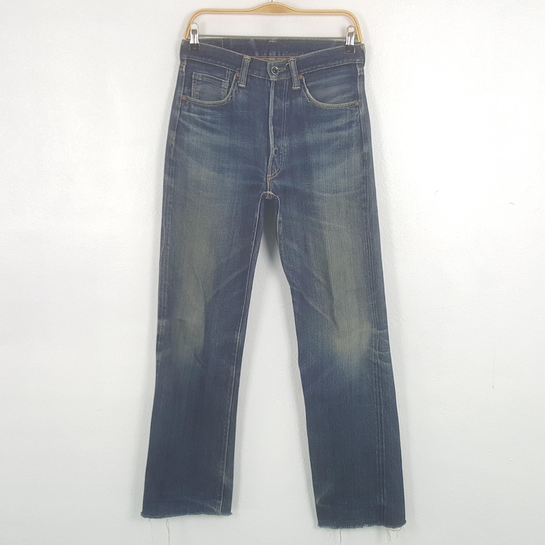 May include: A pair of high-waisted blue denim jeans featuring a faded wash, a button fly closure, and raw, frayed hems at the bottom of the legs. The denim displays natural whiskering patterns at the hips and lighter blue fading at the knees.