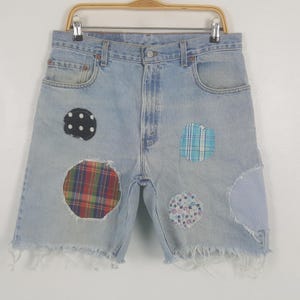 Puede incluir: Shorts vaqueros azul claro con bordes deshilachados y parches de tela coloridos. Los parches incluyen lunares, cuadros escoceses, flores y rayas. Los shorts están colgados de una percha de madera.