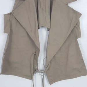 May include: A sleeveless tan vest featuring a wide, draped collar and an open front design. The garment includes two large patch pockets on the lower front panels and thin, matching fabric ties at the waist for an adjustable closure.