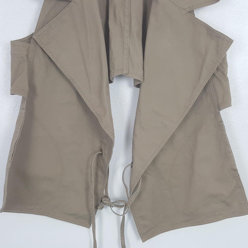 May include: A sleeveless tan vest featuring a wide, draped collar and an open front design. The garment includes two large patch pockets on the lower front panels and thin, matching fabric ties at the waist for an adjustable closure.