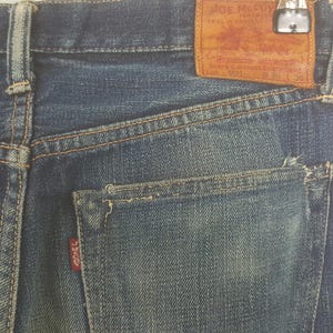 May include: Blue denim jeans featuring a tan leather patch on the back waistband with the text Joe McCoy Real Work Clothes Lot 901 W 29 L 38. The jeans include a back pocket with a small red tab labeled J McCoy and visible orange stitching.