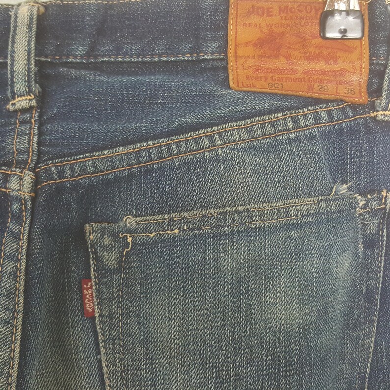May include: Blue denim jeans featuring a tan leather patch on the back waistband with the text Joe McCoy Real Work Clothes Lot 901 W 29 L 38. The jeans include a back pocket with a small red tab labeled J McCoy and visible orange stitching.