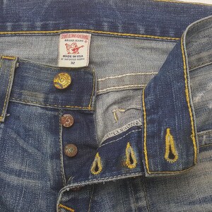May include: Close-up of blue denim jeans with a button fly. The jeans have a faded wash and visible stitching. A label reads "True Religion Brand Jeans" and "Made in USA of Imported Fabric". The size is 32.