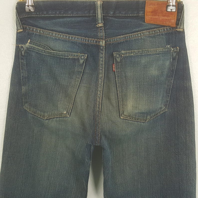 May include: A pair of blue denim jeans featuring a faded wash, two back pockets with frayed edges, a brown leather patch on the waistband, and a small red tab attached to the right back pocket. The jeans are displayed hanging from a metal hanger.