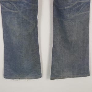 May include: Close-up of the lower portion of a pair of faded blue denim jeans. The jeans have a slight flare at the bottom and show signs of wear and tear, with frayed edges at the hem.
