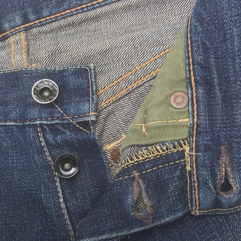 May include: Dark blue denim jeans featuring a button fly closure with two visible black metal buttons and a third button partially hidden. The interior fly flap is lined with olive green herringbone fabric, and the seams feature gold stitching.