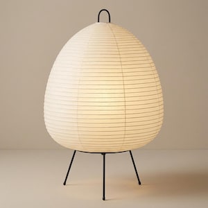 May include: A white paper lantern lamp with a black metal frame. The lamp has an egg-like shape and a black handle at the top. The lamp is supported by three black legs.