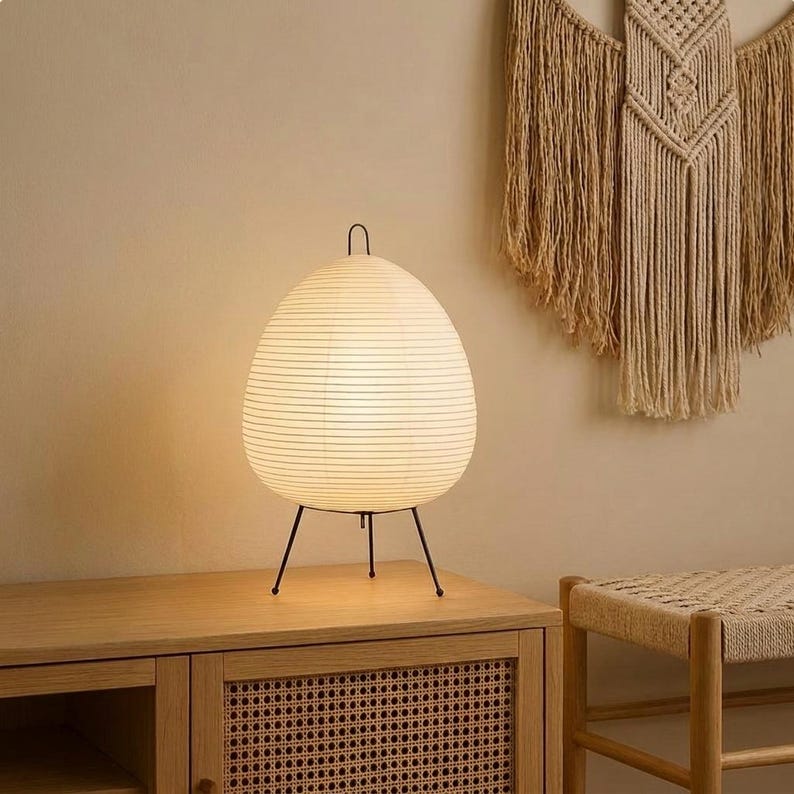 May include: A white paper egg-shaped lamp with a black metal tripod base. The lamp is lit, emitting a warm glow. The lamp sits on a wooden cabinet. The background includes a woven wall hanging and a small bench.