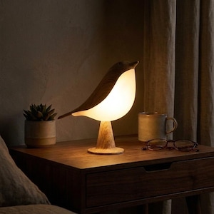 Puede incluir: Lámpara de mesa con forma de pájaro de madera, con cuerpo blanco brillante y base de madera. La lámpara está sobre una mesita de noche de madera oscura, junto a una pequeña planta en maceta, una taza y un par de gafas. La lámpara mide unos 25 cm de alto.