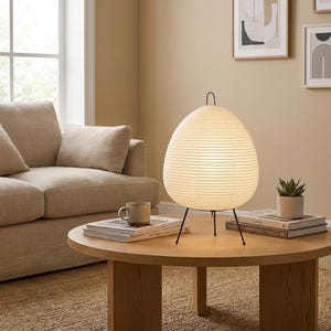 May include: A modern, egg-shaped table lamp with a paper shade and black metal legs. The lamp sits on a round wooden coffee table, alongside books, a mug, and a small potted plant. The room has a neutral color palette.