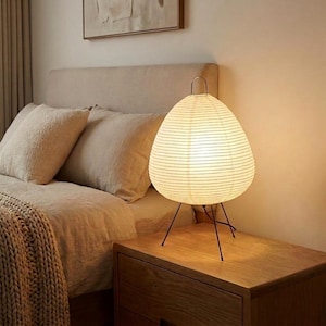 May include: A white paper lantern lamp with a black metal tripod base sits on a wooden nightstand. The lamp has a teardrop shape and is lit from within. The bedroom setting includes a bed with beige bedding and pillows.