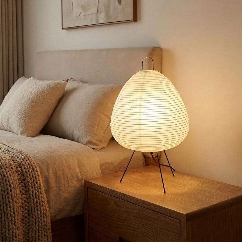 May include: A white paper lantern lamp with a black metal tripod base sits on a wooden nightstand. The lamp has a teardrop shape and is lit from within. The bedroom setting includes a bed with beige bedding and pillows.