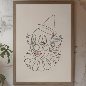 May include: A framed print featuring a minimalist line drawing of a clown's face. The artwork is primarily black, with red lips and blue hair accents. The print is displayed in a light-colored wooden frame, hanging on a textured white wall.