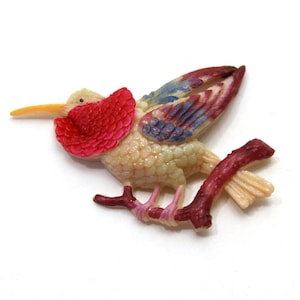May include: A vintage plastic hummingbird brooch with a red, blue, and white painted design. The hummingbird is perched on a red branch.