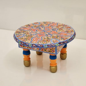 May include: A small, round, wooden table featuring a hand-painted surface with a complex, multi-colored floral and geometric pattern. The table has four short, turned legs painted in alternating bands of bright orange, blue, and gold.