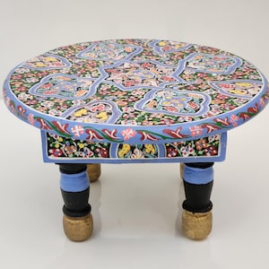 May include: A small round wooden table featuring a vibrant, multi-colored floral pattern across the top and sides. The table is supported by four short legs, each painted with a light blue upper section, a black middle, and a gold base.