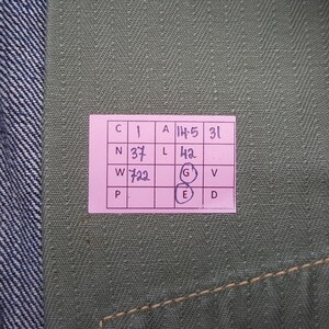 May include: A textured olive green fabric surface featuring a pink rectangular grid label with handwritten black ink text. The grid contains letters and numbers including 1, 14.5, 31, 37, 42, 722, and circled letters G and E, positioned against a blue denim edge.