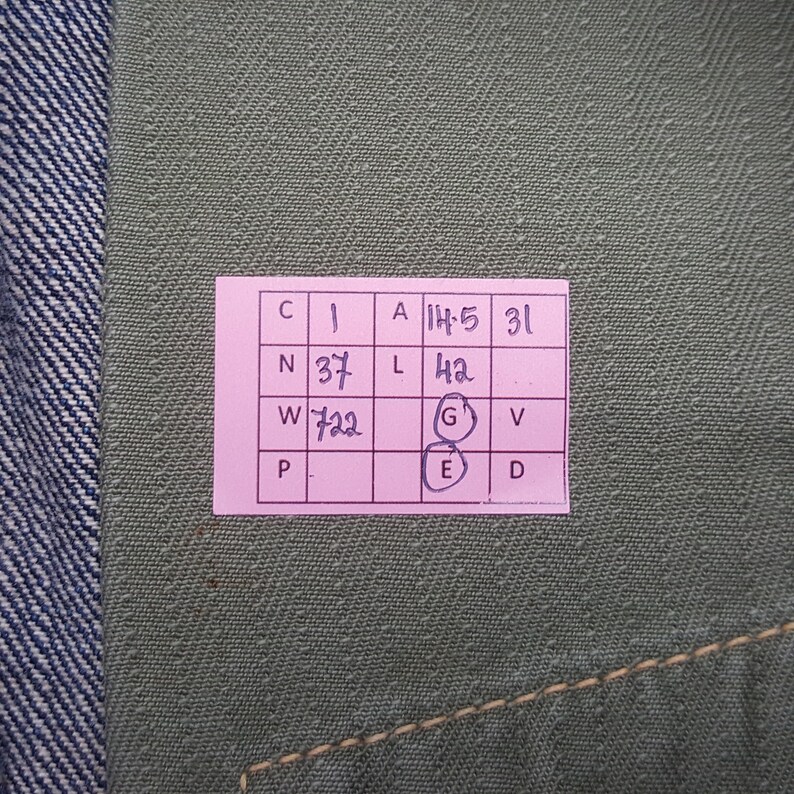 May include: A textured olive green fabric surface featuring a pink rectangular grid label with handwritten black ink text. The grid contains letters and numbers including 1, 14.5, 31, 37, 42, 722, and circled letters G and E, positioned against a blue denim edge.