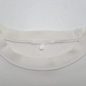 Puede incluir: Primer plano de una sudadera blanca con cuello redondo. Se ven el cuello y los pu&ntilde;os acanalados. Una peque&ntilde;a etiqueta con texto y el n&uacute;mero 9 est&aacute; cosida en el escote.