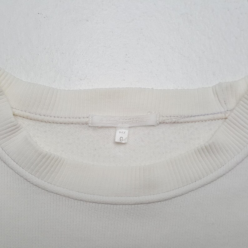 Puede incluir: Primer plano de una sudadera blanca con cuello redondo. Se ven el cuello y los pu&ntilde;os acanalados. Una peque&ntilde;a etiqueta con texto y el n&uacute;mero 9 est&aacute; cosida en el escote.