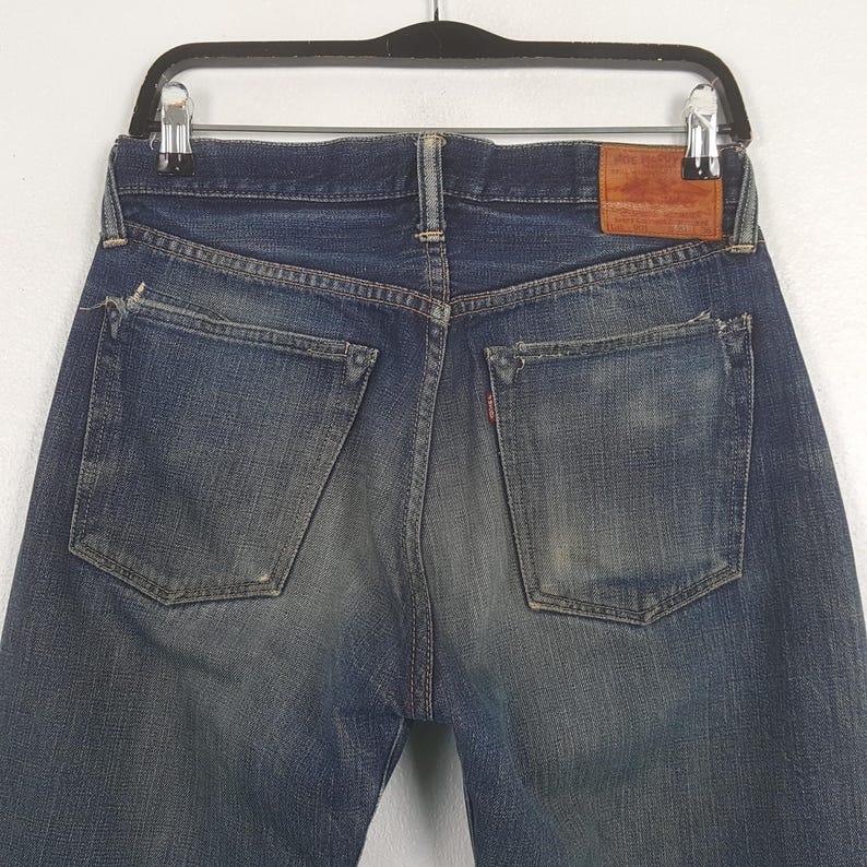 May include: A pair of dark blue denim jeans featuring a faded wash, two back pockets with reinforced corners, a brown leather patch on the waistband, and a small red tab attached to the right pocket. The jeans are displayed on a black hanger.