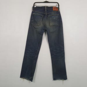 May include: A pair of dark blue denim jeans featuring a faded wash, two back pockets, and a brown leather patch on the waistband. The jeans are displayed on a black hanger and have frayed, raw-edge hems at the bottom of the pant legs.