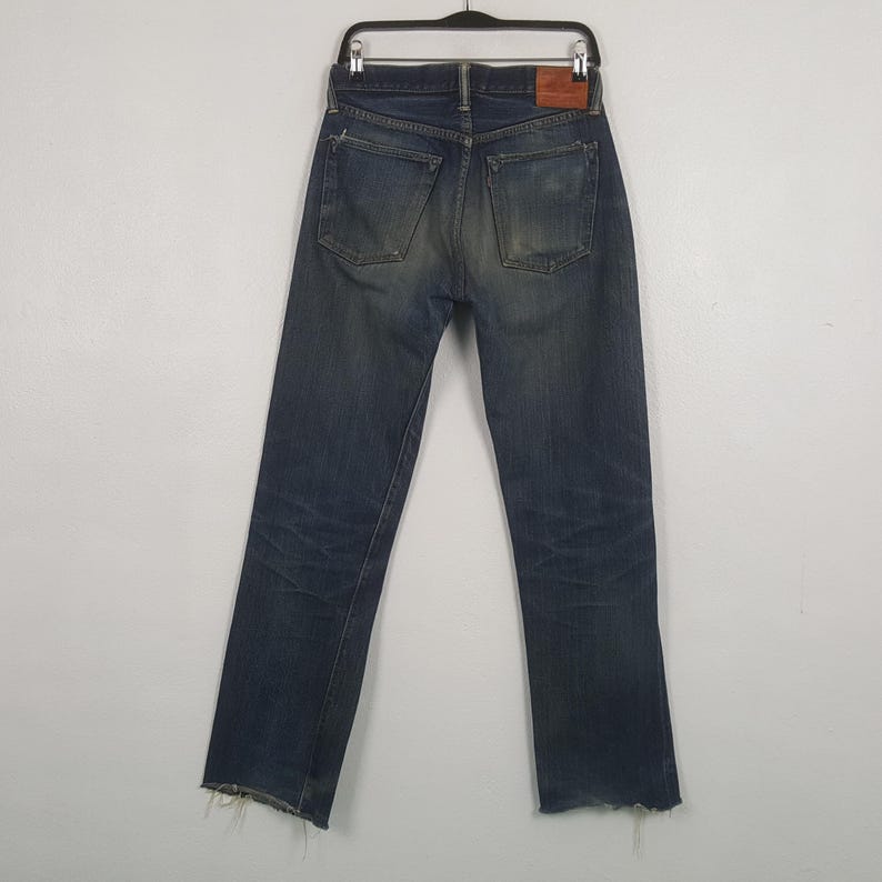 May include: A pair of dark blue denim jeans featuring a faded wash, two back pockets, and a brown leather patch on the waistband. The jeans are displayed on a black hanger and have frayed, raw-edge hems at the bottom of the pant legs.