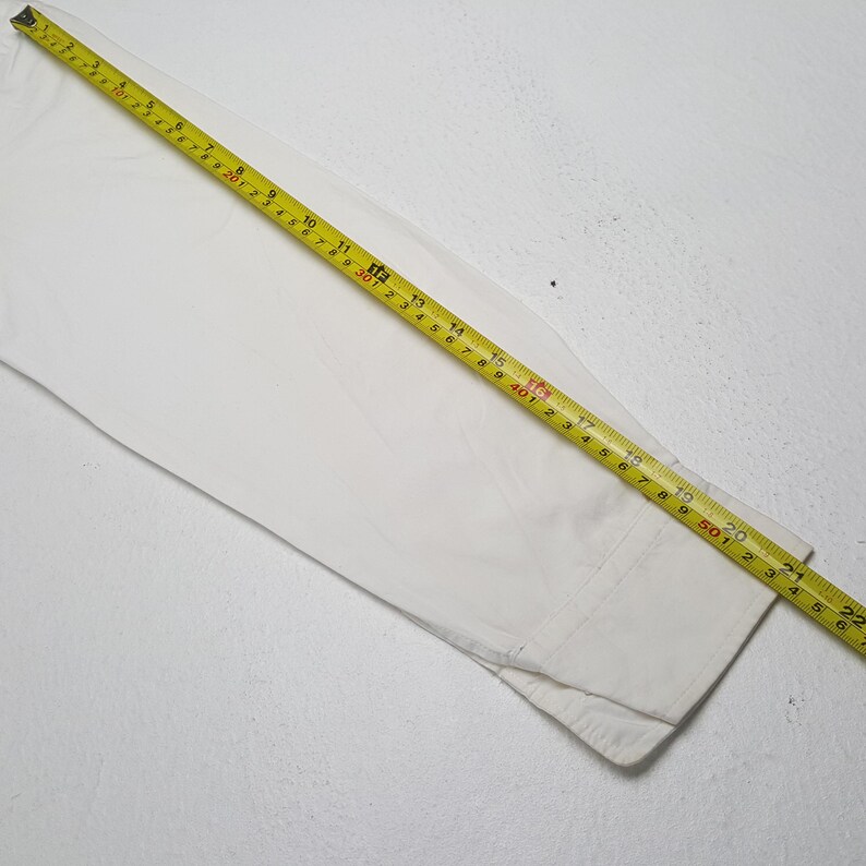 May include: A white long-sleeved shirt with a yellow measuring tape laid across it. The tape shows measurements in inches and feet. The shirt's cuff is visible at the bottom right.