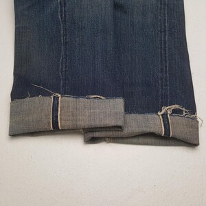 May include: A pair of dark blue denim jeans featuring a straight leg cut with prominent vertical seams running down the front. The cuffs are rolled up to reveal a light grey interior lining with visible frayed edges and a dark blue selvedge detail.