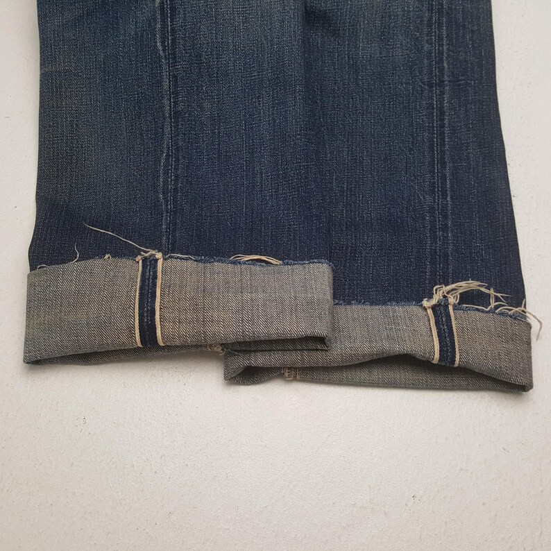 May include: A pair of dark blue denim jeans featuring a straight leg cut with prominent vertical seams running down the front. The cuffs are rolled up to reveal a light grey interior lining with visible frayed edges and a dark blue selvedge detail.