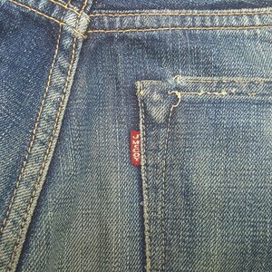 May include: A pair of blue denim jeans featuring a back pocket with thick orange contrast stitching and a small red fabric tab attached to the side of the pocket. The red tab has the white text J MCCOY printed vertically on its surface.