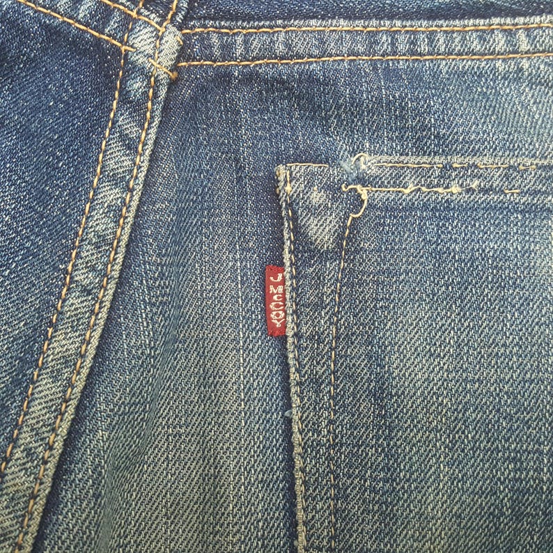 May include: A pair of blue denim jeans featuring a back pocket with thick orange contrast stitching and a small red fabric tab attached to the side of the pocket. The red tab has the white text J MCCOY printed vertically on its surface.