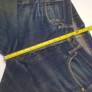 May include: Dark blue denim jeans featuring a classic five-pocket design with copper-toned rivets and a button fly closure. A yellow measuring tape is placed across the waist, showing a measurement of approximately 14.5 inches across the top.