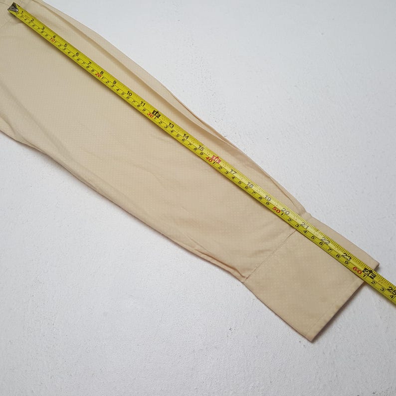 Pode incluir: Uma manga comprida de uma camisa bege est&aacute; estendida com uma fita m&eacute;trica amarela colocada diagonalmente sobre ela, mostrando um comprimento de aproximadamente 63,5 cm da costura do ombro at&eacute; a borda do punho na manga.