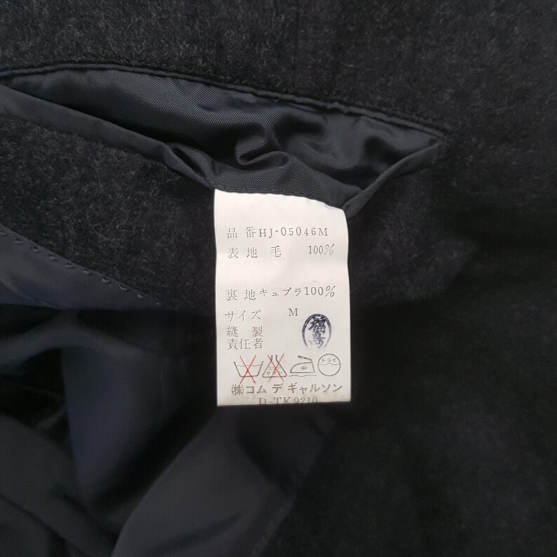 May include: Close-up of a dark gray wool garment tag with Japanese text. The tag includes the product code HJ-05046M, fabric composition, size M, and care symbols. The garment appears to be a jacket or coat.