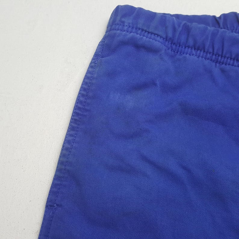Puede incluir: Primer plano de una secci&oacute;n de tela azul, posiblemente pantalones cortos o pantalones. La tela tiene una textura visible y una costura cosida a lo largo del borde. La cintura es el&aacute;stica. El fondo es una superficie blanca lisa.